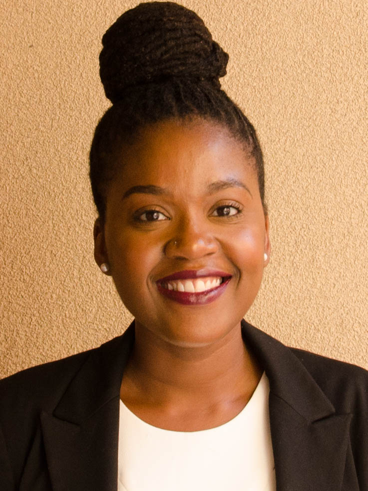 Headshot of a woman in a white t-shirt and black blazer.  She has her hair up in a top knot and has a bright smile.