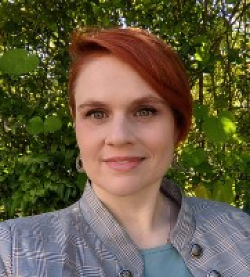 Gretchen is a red-headed woman smiling at the camera; she is wearing a grey-blue suit jacket.