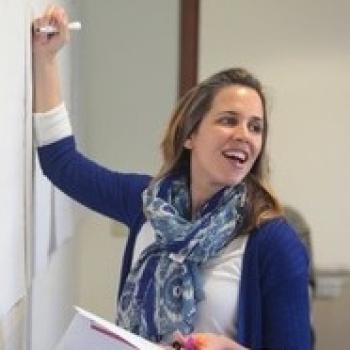 Woman with auburn hair writing on a whiteboard, wearing a dark blue cardigan, white blouse, and a blue floral patterned scarf.